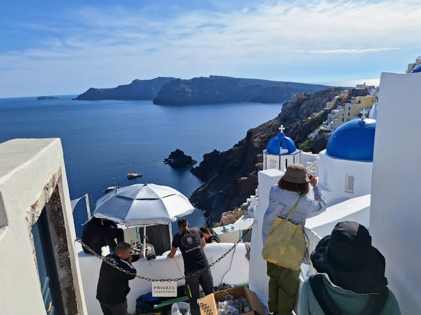 Crowded viewpoint in Oia Santorini with tourists taking pictures of the blue domes while there's a busy construction crew and metal chain to bar entry.