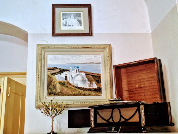 Framed painting of a blue-domed Santorini chapel above an antique wind-up gramophone and a brass olive-tree ornament in a whitewashed room you can visit in Santorini without feeling like a tourist