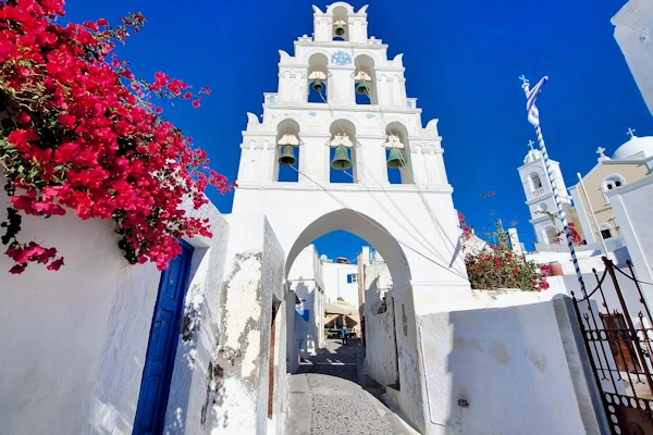 Whitewashed Cycladic church belfry with a stone archway with pink bougainvillea and a cobbled lane in Megalochori. Santorini