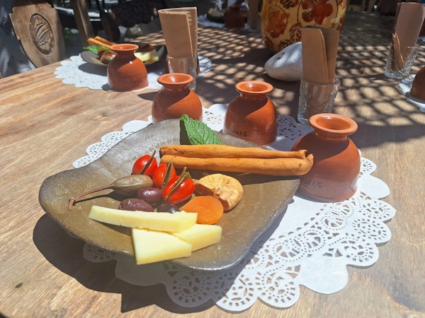 Stone plate of Santorian mezze — graviera cheese, olives, cherry tomatoes, dried fig and apricot — beside handmade terracotta Symposion cups in dappled light.